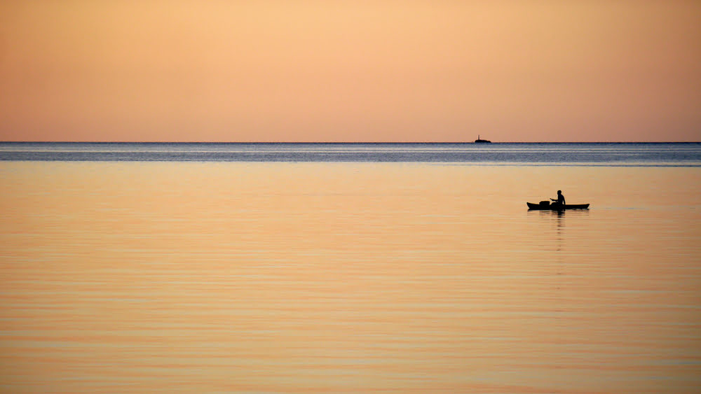 Ein Fischer in einem kleinen Boot auf dem ruhigen Wasser des Malawisees bei Sonnenuntergang mit spiegelnder Wasseroberfläche und weitem Horizont.