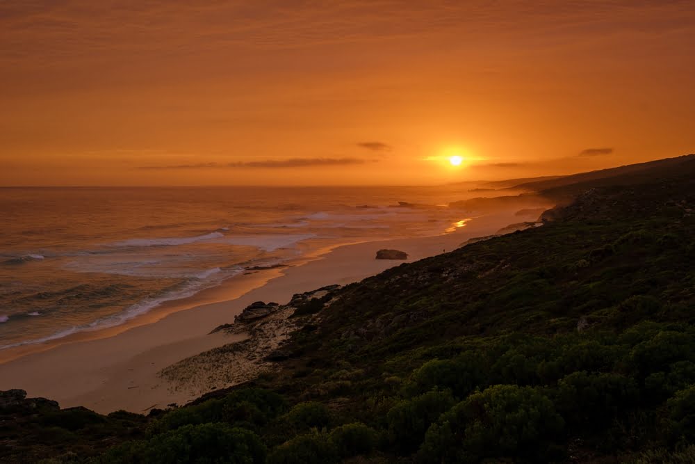 Sonnenuntergang an der Küste der Garden Route in Südafrika mit langen Sandstränden, rauem Ozean und grünen Hügeln.